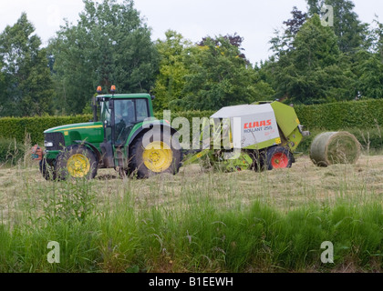 Grüne John Deere Tractor Pulling eine Rundballenpresse während der Heuernte in einem Feld von Cheshire Stockfoto