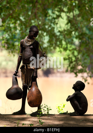 Eine Karo-Frau hält zu plaudern, ein kleiner Junge vor dem Befüllen ihre große Kürbisse mit Wasser aus dem Omo-Fluss. Stockfoto