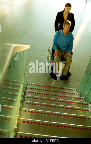 Männlicher Patient sitzt in einem Rollstuhl und eine Mitte erwachsenen Frau stand neben ihm in der Nähe eine Treppe Stockfoto