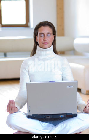 junge attraktive Frau praktizieren Yoga mit einem Notebook auf dem Schoß sitzen auf dem Boden im Wohnzimmer Stockfoto