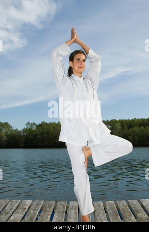 junge hübsche Frau auf einem Holzsteg am See, Baum, Vrikshasana Yoga praktizieren Stockfoto