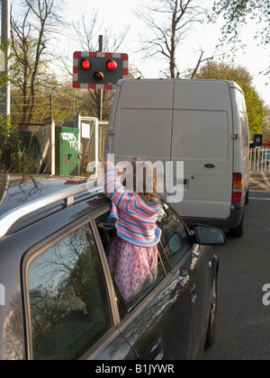 Fahrzeuge warten auf Eisenbahn Bahnübergang Barriere für Zug übergeben, & kleines Kind hängt gefährlich aus einem Autofenster zu beobachten. Stockfoto