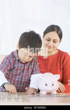 Junge Stapel von Münzen in der Nähe ein Sparschwein mit seiner Mutter, die neben ihm sitzen Stockfoto