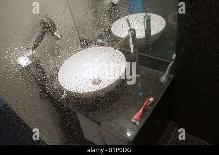 Ein modernes Badezimmer mit einem Waschbecken, das durch eine mit Wassertropfen bedeckte Glasscheibe blickt. Stockfoto