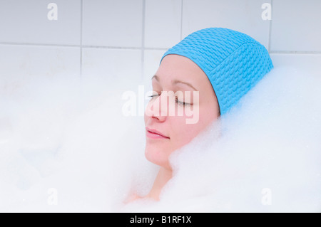 Hübsche junge Frau unter Bad in der Badewanne und entspannend, Badekappe blau Stockfoto
