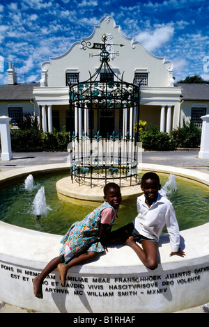 Zwei glückliche afrikanische Kinder sitzen am Brunnen vor dem Rathaus von Franschhoek in der Nähe von Stellenbosch, Western Cape, So Stockfoto