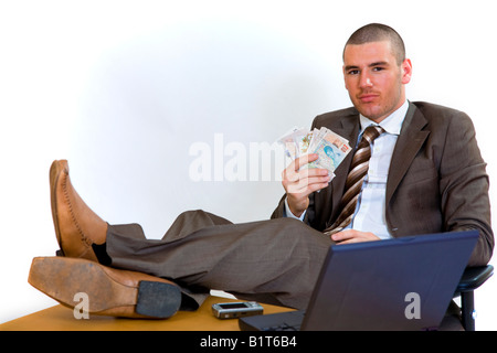 Junger Geschäftsmann mit Füßen auf seinem Schreibtisch und hält ein Bündel Geld. Stockfoto