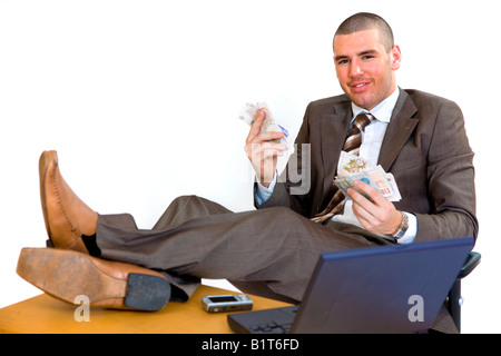 Junger Geschäftsmann mit Füßen auf seinem Schreibtisch und hält ein Bündel Geld. Stockfoto