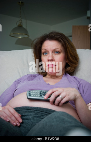 Schwangere Frau sitzend auf einem Stuhl, halten die Fernbedienung wie sie Modell veröffentlicht fernsieht Stockfoto