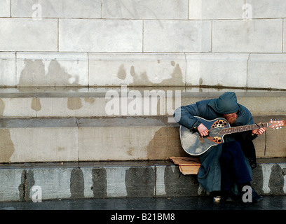 Ein Obdachloser Mann sitzt beim Musizieren auf Passanten auf den Straßen von Dublin, in der Hoffnung, etwas Kleingeld zu gewinnen. Stockfoto