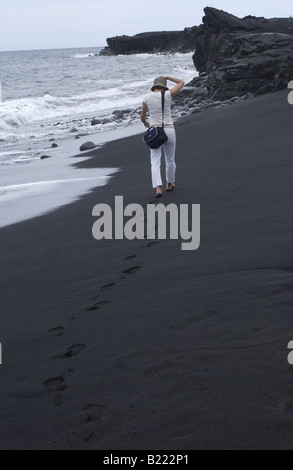 Eine Frau mit einer Kameratasche Fuß einen schwarzen Sandstrand auf der Insel Hawaii. Stockfoto