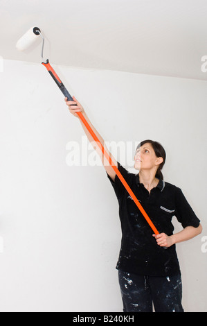 Eine weiße Frau mit langen braunen Haaren malt eine Decke mit einer Malerwalze mit langem Griff und weißer Emulsionsfarbe. Stockfoto