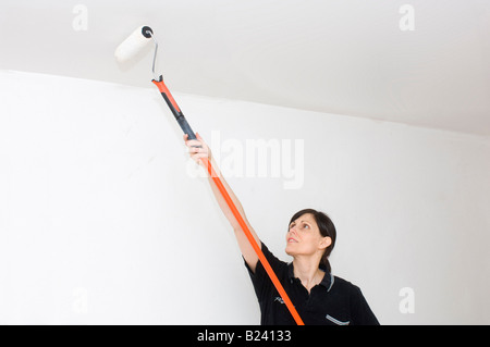 Eine weiße Frau mit langen braunen Haaren malt eine Decke mit einer Malerwalze mit langem Griff und weißer Emulsionsfarbe. Stockfoto