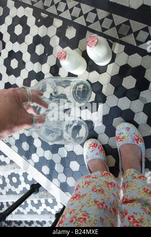 Das Leergut auf eine britische Haustür eine Frau im geblümten Pyjama Außerbetriebnahme legt leer Milkbottles auf den Schritt für das recycling Stockfoto