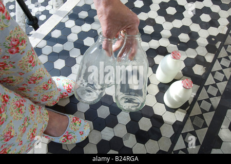 Das Leergut auf eine britische Haustür eine Frau im geblümten Pyjama Außerbetriebnahme legt leer Milkbottles auf den Schritt für das recycling Stockfoto