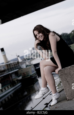 süße Braunhaarige Frau in einem kurzen schwarzen Kleid, sitzt an der harbourl Stockfoto