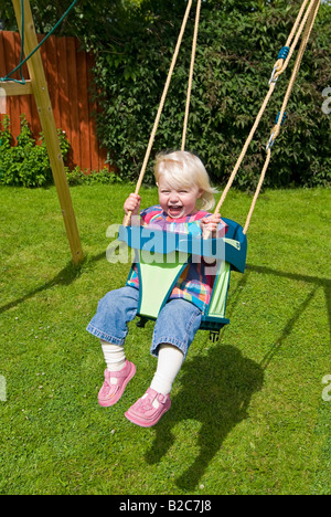 Vertikale Porträt eines 18 Monate alten kleinen Mädchen mit viel Spaß spielen auf ihrer Schaukel draußen im Garten an einem sonnigen Tag Stockfoto