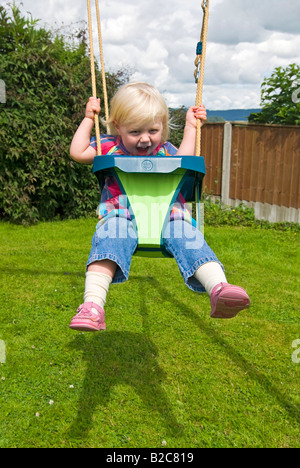 Vertikale Porträt eines 18 Monate alten kleinen Mädchen mit viel Spaß spielen auf ihrer Schaukel draußen im Garten an einem sonnigen Tag Stockfoto