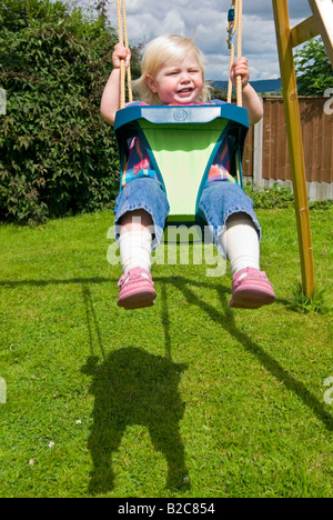 Vertikale Porträt eines 18 Monate alten kleinen Mädchen mit viel Spaß spielen auf ihrer Schaukel draußen im Garten an einem sonnigen Tag Stockfoto