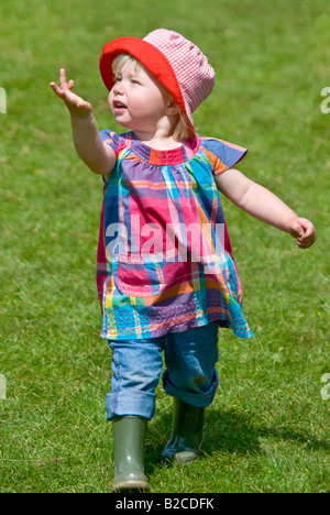 Vertikale Porträt eines 18 Monate alten kleinen Mädchen mit einen hellen roten Sonnenhut auf laufen im grünen an einem sonnigen Tag. Stockfoto