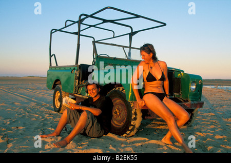 Touristen am Strand eine Frau in einem Bikini und einem Mann lehnte sich gegen einen alten Safari-Fahrzeug, ein Buch zu lesen. Stockfoto