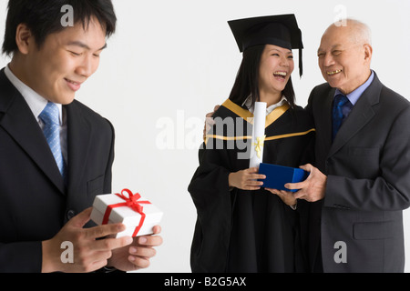 Junge weibliche Absolventin erhält ein Geschenk von ihrem Vater und ein junger Mann hält ein Geschenk Stockfoto