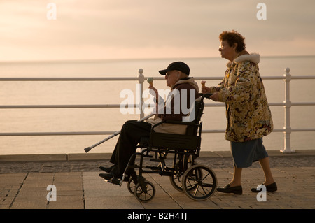 Ältere Frau Pfleger schob ihren behinderten Mann im Rollstuhl beide essen Eis am Meer Sommerabend, Wales UK Stockfoto