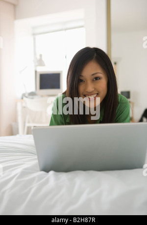 Porträt einer jungen Frau liegend auf dem Bett vor einem Laptop und lächelnd Stockfoto