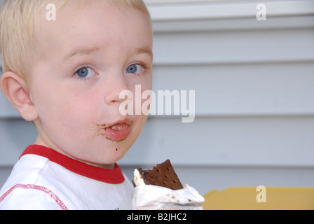 Ein Kleinkind junge isst eine Eis-Sandwich an eine Kinder-Picknick-Tisch draußen in einer Sommernacht. Stockfoto