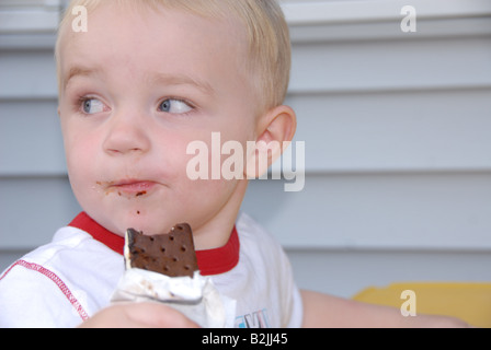 Ein Kleinkind junge isst eine Eis-Sandwich an eine Kinder-Picknick-Tisch draußen in einer Sommernacht. Stockfoto