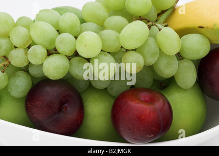 Nahaufnahme einer weißen Schale mit Früchten, die frische rohe Pflaumen, grüne Trauben, rote und grüne Äpfel und eine Banane enthält Stockfoto