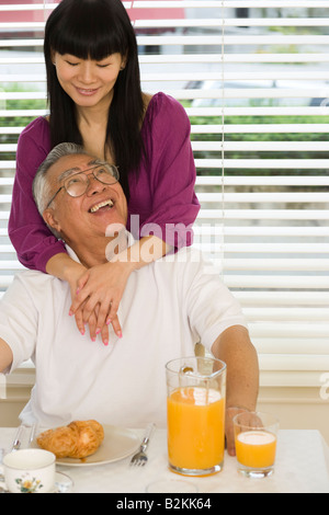 Mitte Erwachsene Frau umarmt ihr Vater von hinten einen Frühstückstisch Stockfoto
