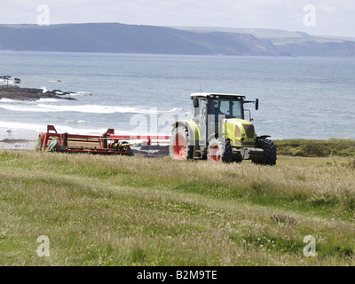 Landwirt einen Traktor schneiden Gras in einem Feld neben dem Meer, ein Beispiel der küstennahen Landwirtschaft, Bude, Cornwall, UK Stockfoto