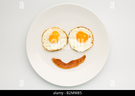 Zwei Spiegeleier und ein kleines knuspriges Speckchen auf einem weißen Teller in Form eines Smiley-Gesichts. Flach verlegt. Stockfoto