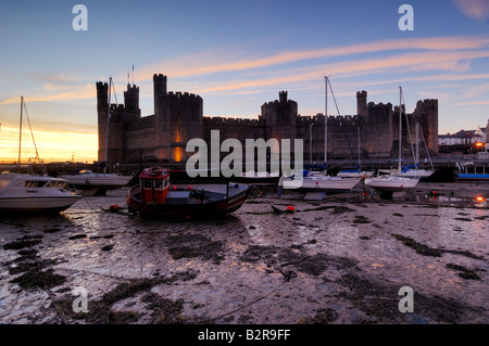 Caernarfon Castle an der Küste von North Wales beleuchtet in der Nacht mit den Booten in der Mündung auf die Ebbe gestrandet Stockfoto