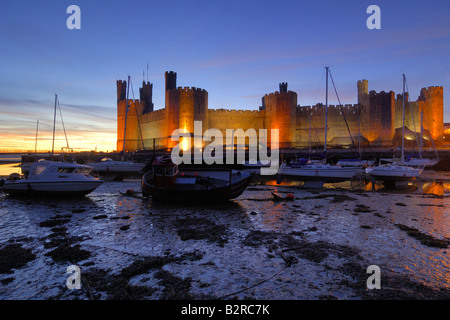 Caernarfon Castle an der Küste von North Wales beleuchtet in der Nacht mit den Booten in der Mündung auf die Ebbe gestrandet Stockfoto