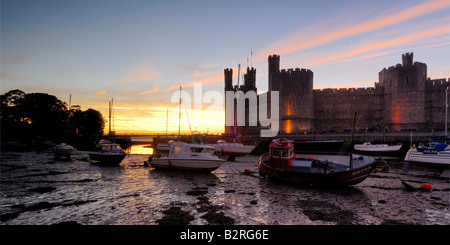 Caernarfon Castle an der Küste von North Wales beleuchtet in der Nacht mit den Booten in der Mündung auf die Ebbe gestrandet Stockfoto