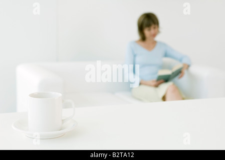 Frau liest Buch auf Sofa, Schwerpunkt Kaffeetasse im Vordergrund Stockfoto