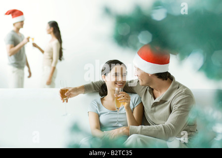 Paar auf Sofa sitzen, trinken Champagner, man trägt Weihnachtsmütze Stockfoto