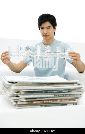 Junger Mann mit gestapelten Zeitungen halten Baum Formen schneiden aus Papier, Blick in die Kamera Stockfoto