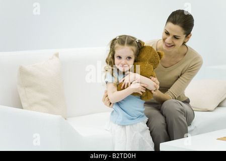 Kleines Mädchen hält ausgestopfte Bär, Frau sitzt auf der Couch, sanft umarmt kleine Mädchen Stockfoto