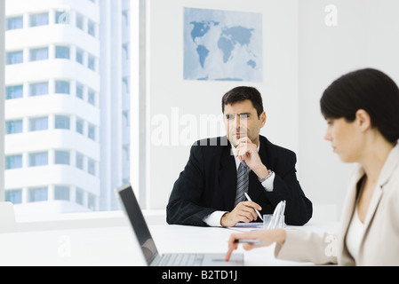 Exekutive sitzen im Büro mit der Hand unter Kinn, Kollegin mit Laptop im Vordergrund Stockfoto