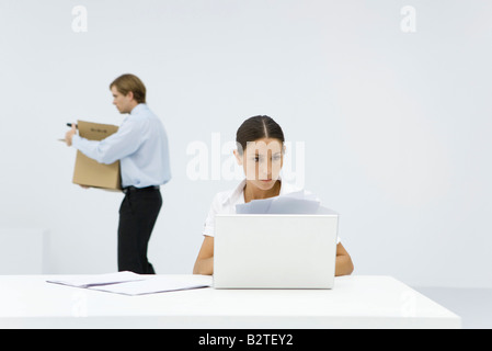 Berufstätige Frau überprüfen von Dokumenten am Schreibtisch hinter Laptop, Kollege tragen Kasten im Hintergrund Stockfoto