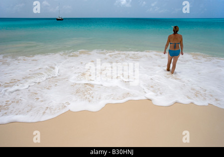 Eine Frau watet in der sanften Brandung 11 Mile Beach auf Barbuda s West Coast, die ihr Segelboot auf dem Anker direkt vor der Küste schwingt Stockfoto