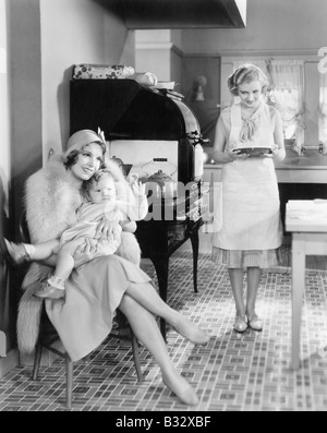 Elegante Frau mit ihrem Baby in der Küche sitzen, während ein Freund eine Torte bringt Stockfoto