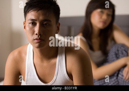 Ein asiatischer Mann sieht unglücklich mit seinem Partner (out of Focus) hinter ihm auf dem Bett sitzend Stockfoto