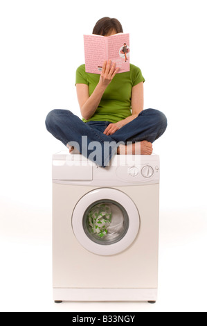 Weiße Frau mit dunklen Haaren, mit grünem Oberteil und blauer Jeans. Sie sitzt kreuzbeinige Beine auf einer Haushaltswaschmaschine und liest ein Buch. Stockfoto