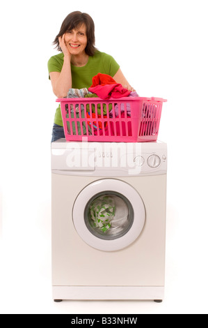 Lächelnde junge weiße Frau, die ein grünes T-Shirt trägt und sich auf einen pinkfarbenen Wäschekorb voller Kleidung lehnt, der sich auf einer Waschmaschine befindet Stockfoto