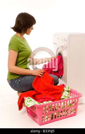 Junge weiße Frau mit einem grünen T-Shirt und Jeans, die Haushaltswaschmaschine mit bunten Kleidern aus dem Wäschekorb beladen. Stockfoto