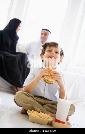 Junge mit Fast Food im Wohnzimmer lachend mit Eltern im Hintergrund (hohe Schlüssel/selektiven Fokus) Stockfoto
